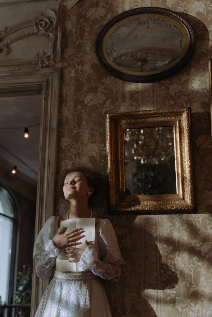 A beautiful lady in a white dress leaning back against a wall with a letter in her hands looking like she is dreaming.