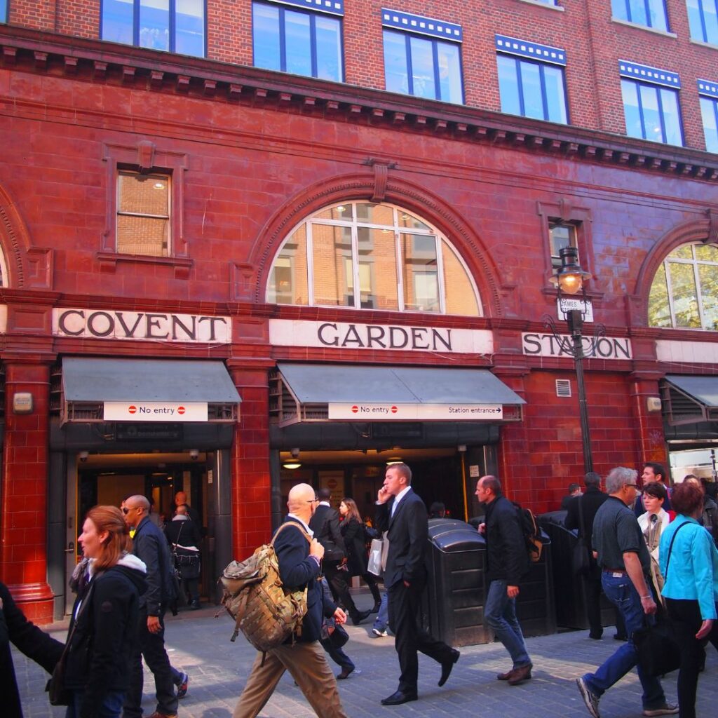 The Covent garden station that is a red building in a trendy neighborhood in London and there are a lot of people walking in front of it.