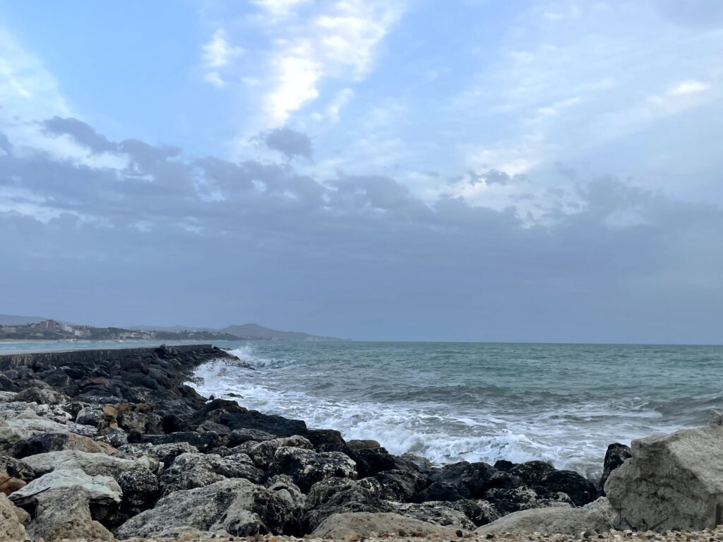 Waves breaking on a rocky breakwater along the Sicilian coast under a grey sky.
