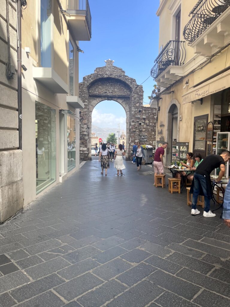 A historic stone archway gate at the end of Corso Umberto, Taormina's main pedestrian street, lined with cafes and shops, Sicily.