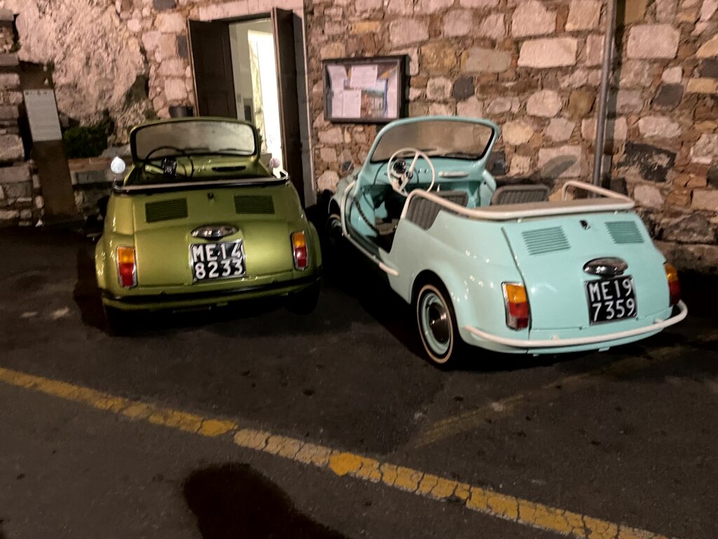 Two classic Fiat 500 cars parked by a stone wall at night in Sicily.